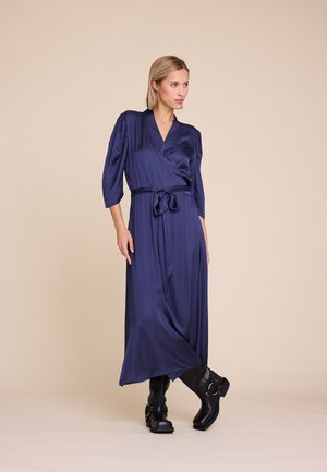 Kvinne iført en lang marineblå omslagskjole med trekvartlange ermer og bundet midje, satt sammen med svarte støvler til midtkalv, mot en beige bakgrunn.