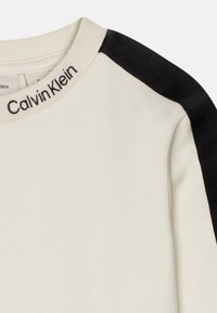 Sudadera de color crema con acentos negros en los hombros, con cuello acanalado y logo bordado de "Calvin Klein". Textura suave.