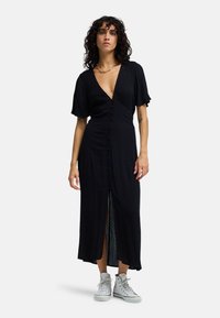 Μαύρο φόρεμα μέχρι το γόνατο με κοντά, φαρδιά μανίκια· διαθέτει βαθύ V-λαιμόκοψη, κουμπιά μπροστά και σκίσιμο στο πλάι· συνδυασμένο με λευκά αθλητικά παπούτσια.