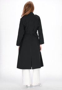 Zwarte trenchcoat met riem, lange mouwen, achtersplit en een gladde textuur. Draagt over witte wijde broekspijpen en gestreepte schoenen.