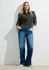 Camicia button-up grigia scura a maniche lunghe, abbinata a jeans a zampa d'elefante blu. Il modello è in piedi con le mani in tasca su uno sfondo chiaro.
