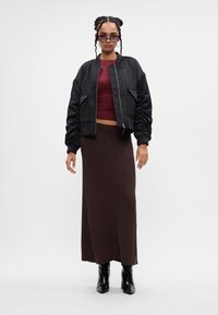 Kobieta z warkoczami, nosząca okulary przeciwsłoneczne, czarną bomberkę, bordowy top, długą brązową spódnicę i czarne botki na obcasie, stoi na białym tle.