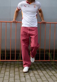 Vit, kortärmad sporttröja med röda och gula accenter, kombinerad med löst sittande rosa byxor och vita sneakers. Orange räcke i bakgrunden.