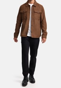 Homme portant une veste à boutons marron, un t-shirt blanc, un pantalon noir et des bottes noires à lacets, se tenant devant un fond uni.