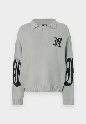 Jersey tejido de color gris claro con dobladillo y cuello acanalados. Presenta detalles de logo en negro en el pecho y las mangas. Textura suave.