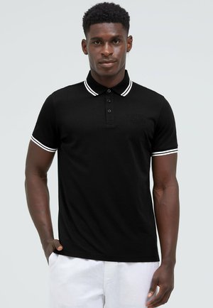 Man standing with one hand in pocket wearing a black polo shirt with white striped collar and sleeves, and white pants.