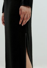 Schwarzes Kleid aus strukturiertem Stoff, mit einem seitlichen Schlitz und kontrastierenden weißen Nähten. Nahaufnahme einer Hand, die auf dem Kleid ruht.
