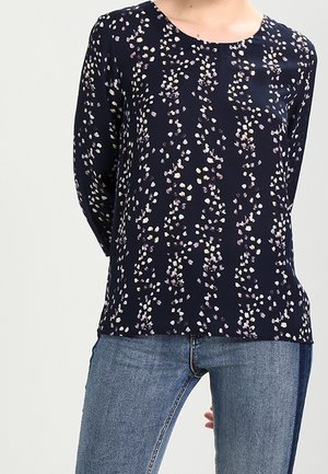 Žena v tmavě modré blůze s dlouhým rukávem, zdobené bílými a fialovými květinovými puntíky, v kombinaci se světle modrými džínami, stojící před jednobarevným pozadím.