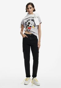 T-shirt graphique gris avec Mickey Mouse, associé à un pantalon cargo noir et des baskets blanches avec des lacets jaunes. Le modèle pose avec confiance.
