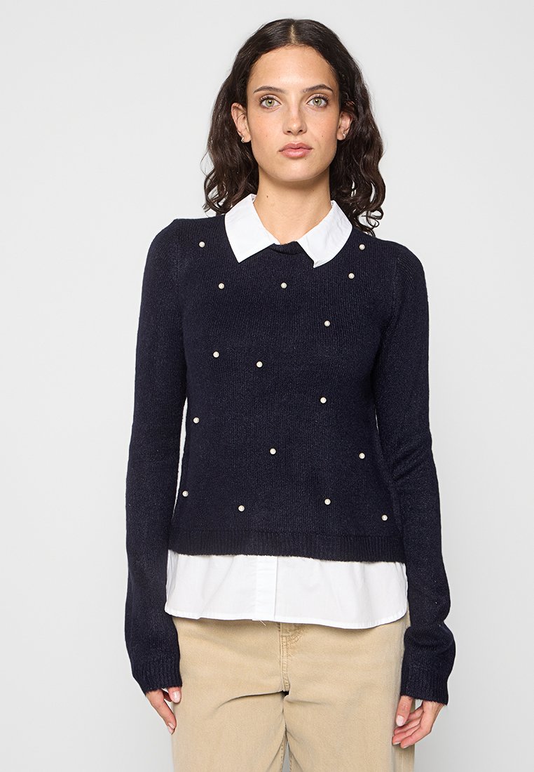 Pull en maille marine avec accents de perles, col rond et manches longues, porté sur une chemise blanche à col. Tissu texturé avec ourlet côtelé.