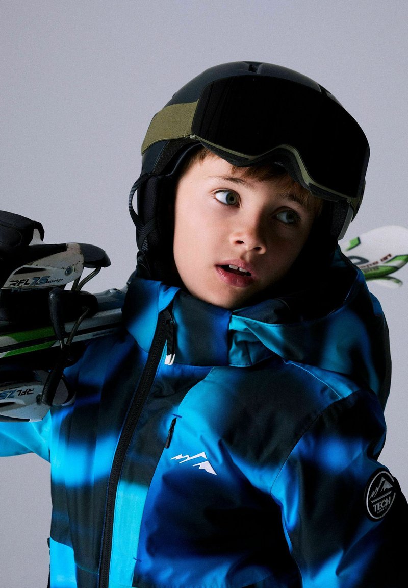Veste de ski bleue et noire avec fermeture éclair et logo de montagne, portée avec un casque noir et des lunettes de ski, tenant des skis.