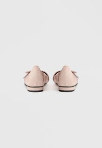 Sapatilhas em camurça rosa com detalhe de laço no calcanhar, apresentando sola baixa preta e design de bico arredondado. Superfície texturizada que realça o interesse visual.