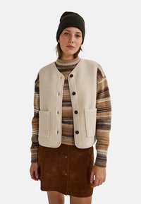 Jeune femme portant un bonnet noir, un gilet en polaire beige avec des boutons, un pull rayé marron et une jupe en suède marron avec des boutons, se tenant devant un fond blanc.