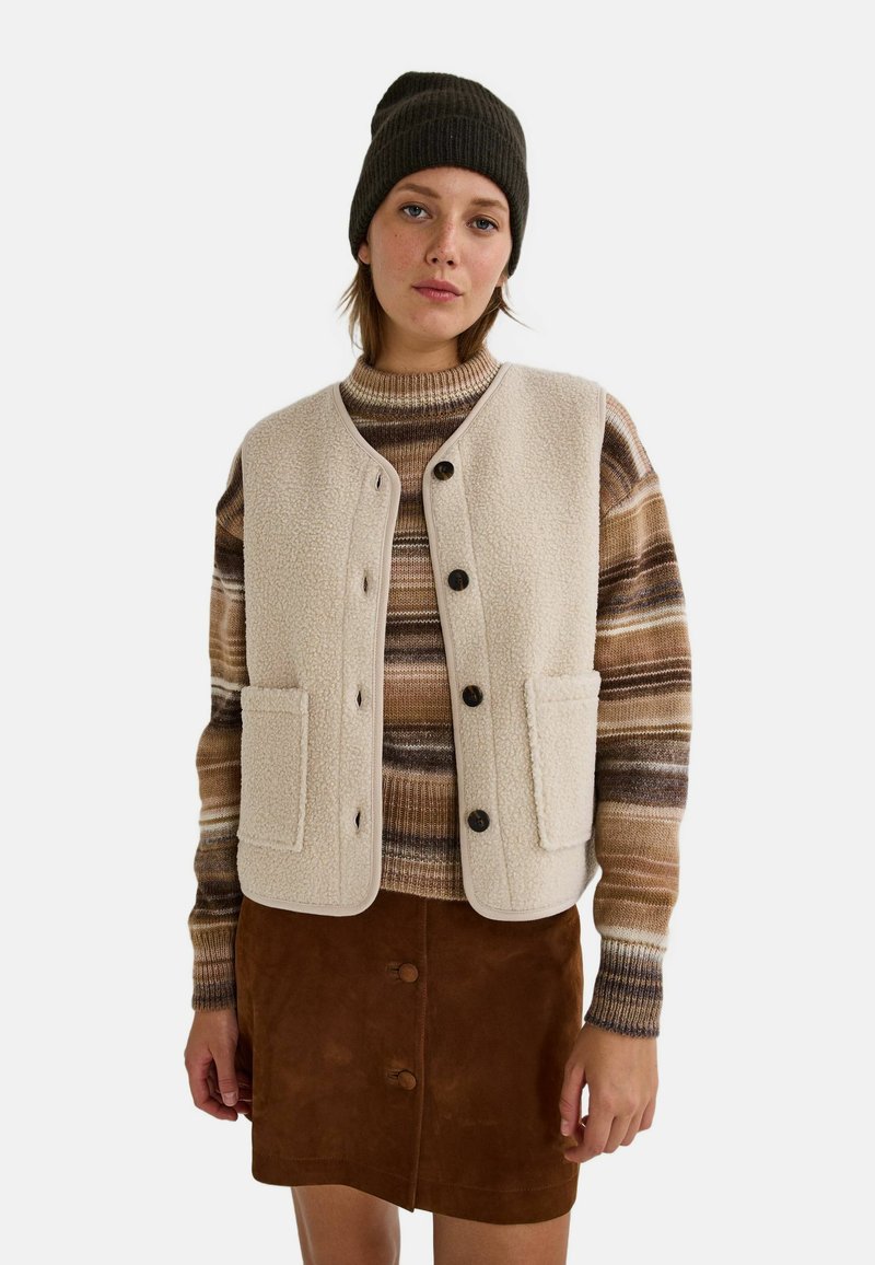 Jeune femme portant un bonnet noir, un gilet en polaire beige avec des boutons, un pull rayé marron et une jupe en suède marron avec des boutons, se tenant devant un fond blanc.