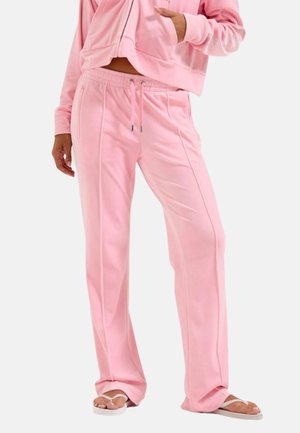 Vrouw met losse roze velour joggingbroek en bijpassende hoodie, met één hand in de zak, staand op witte flipflops.