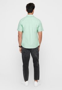 Uomo in piedi di spalle, che indossa una camicia a maniche corte verde chiaro, jeans neri, sneakers bianche e un orologio da polso nero al polso sinistro.