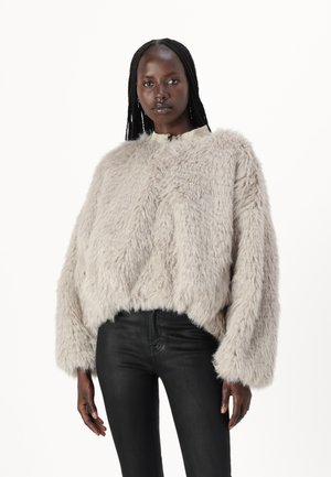 Pull en fausse fourrure beige clair avec une coupe carrée et un col haut. Présente des manches larges et un extérieur texturé. Associé à un pantalon noir.