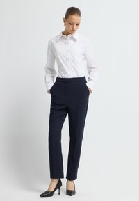 Bílá košile s knoflíky a kapsou, doplněná navy kalhotami na míru. Černé vysoké podpatky s texturovaným vzorem. Jednoduché pozadí.