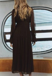 Un vestido largo de color marrón oscuro hecho de una tela fluida con detalles plisados y mangas largas transparentes, que cuenta con un corsé ajustado y una falda acampanada.