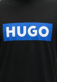 Zwart shirt met een gedurfd blauw rechthoekig logo dat het woord "HUGO" in grote witte hoofdletters in het midden van de borst toont.