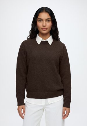 Femme aux longs cheveux foncés portant un pull marron foncé par-dessus une chemise blanche à col et un pantalon blanc, debout devant un fond clair uni.