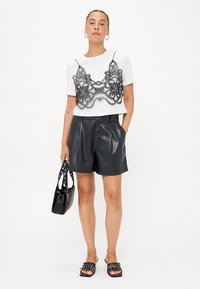 Top corto blanco con detalles de encaje negro, pantalones cortos de faux cuero negro, sandalias negras tipo slide y un bolso negro con tachuelas.