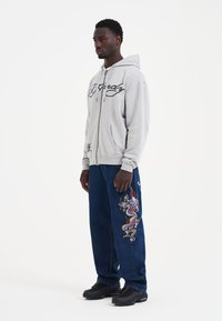 Gray zip-up hoodie with logo, paired with baggy dark blue jeans featuring a colorful dragon embroidery, and black sneakers.