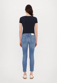 Femme debout de dos, portant un t-shirt bleu marine, un jean skinny bleu clair et des baskets blanches, sur un fond blanc uni.