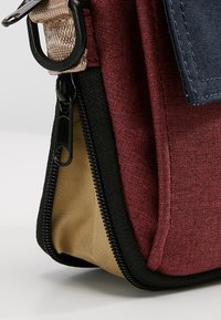 Burgundy tygväska med svart dragkedja, beige botten och marinblå detaljer. Har en strukturerad yta och metallbeslag på remmen.