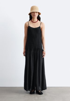 Femme debout portant une longue robe plissée noire, un chapeau beige type bob, un collier de perles rouges, des bracelets en argent et des ballerines noires sur un sol gris.