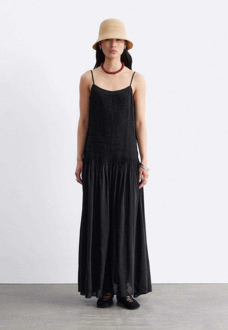 Femme debout portant une longue robe plissée noire, un chapeau beige type bob, un collier de perles rouges, des bracelets en argent et des ballerines noires sur un sol gris.