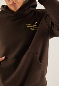 Felpa marrone con tasca frontale, cappuccio regolabile con lacci e testo stampato giallo che recita "PIER ONE OUTDOOR SUPPLY" insieme a una coordinata.