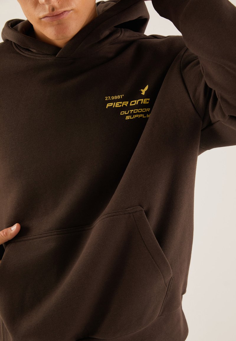 Sudadera marrón con un bolsillo frontal, capucha con cordón y texto impreso en amarillo que dice "PIER ONE OUTDOOR SUPPLY" y unas coordenadas.