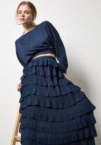 Marineblaues Crop-Top mit langen Ärmeln, kombiniert mit einem plissierten Maxirock mit geschichteten Rüschen, die eine strukturierte, fließende Silhouette erzeugen.