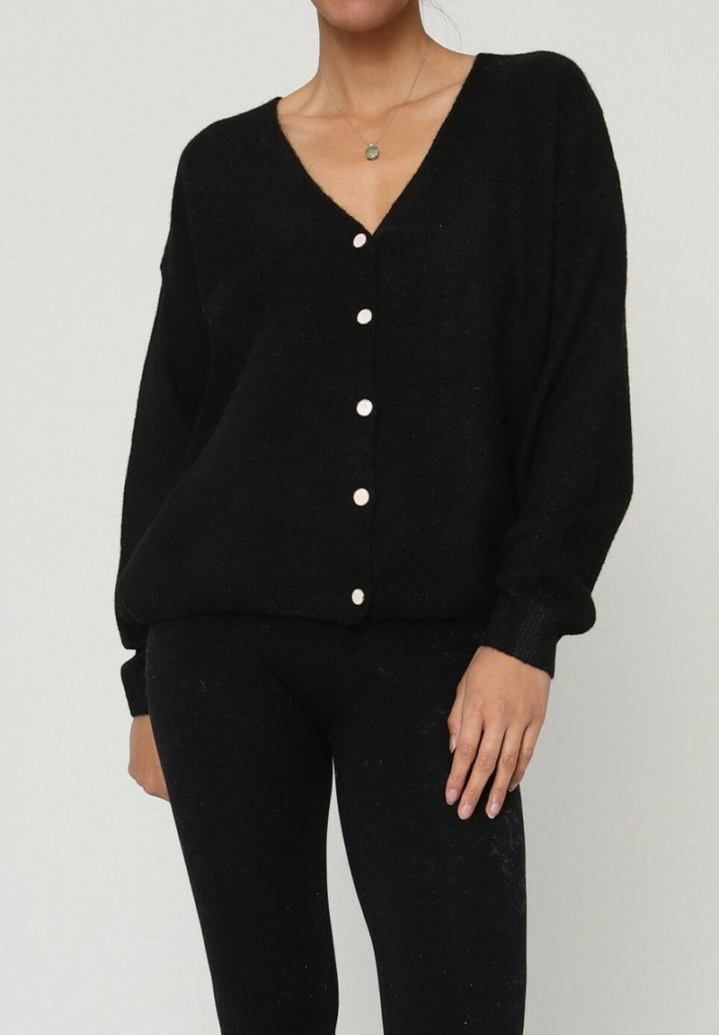 Cardigan noir surdimensionné avec un col en V, doté de gros boutons blancs et d'une texture douce. Porté par-dessus des leggings noirs ajustés.