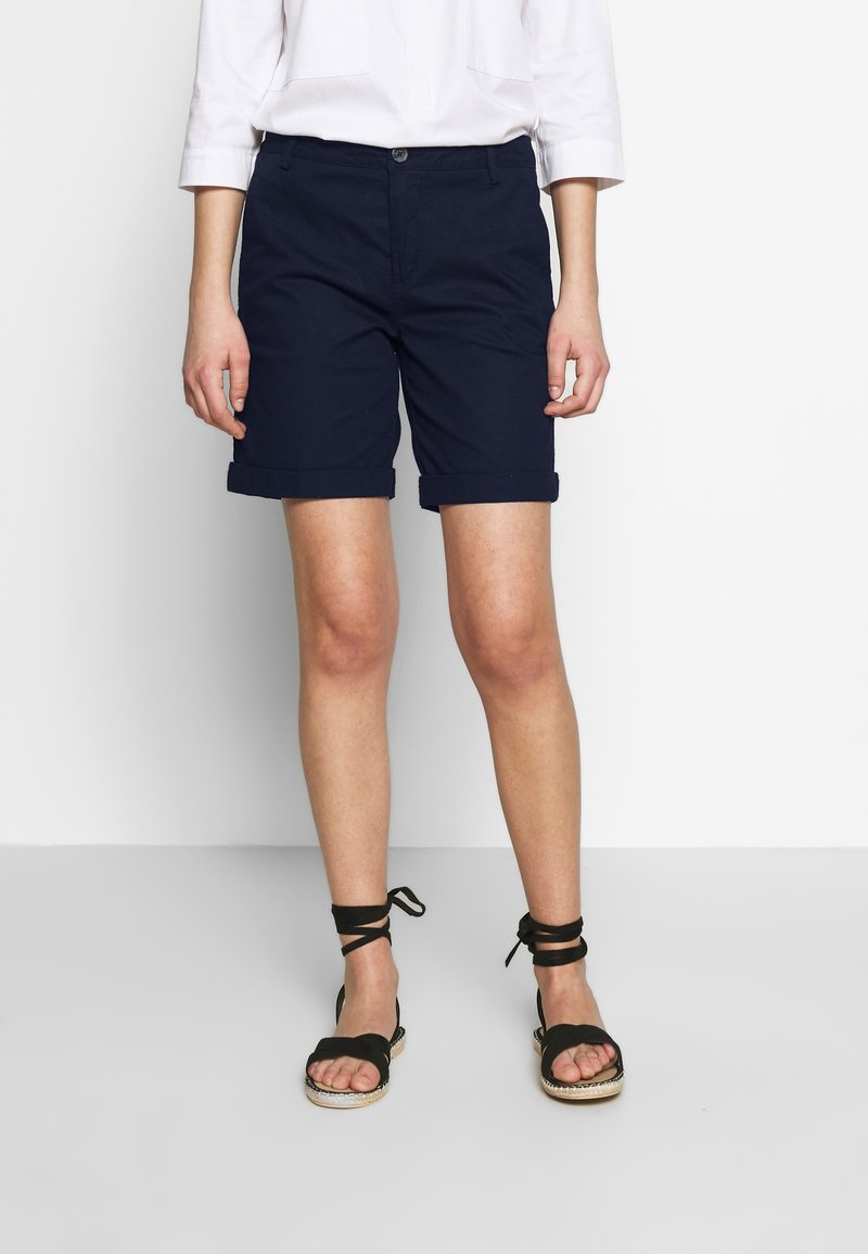 Marina blå bomullsshorts med upprullade fållar, framknäppning och sidofickor, tillsammans med svarta sandaler med remmar.