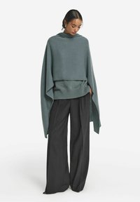 Överdimensionerad teal poncho med huva och ribbad fåll, lager på lager över en mörkgrå tröja, parat med vida svarta byxor.