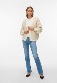 Bēša adīta kardigāna ar teksturētu virsmu, plati piedurknes un V-veida izgriezumu, kopā ar gaiši zilām taisna griezuma džinsām.