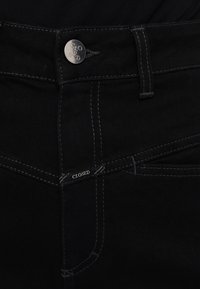 Jean en denim noir avec coutures blanches, avec un bouton métallique et une petite étiquette, tous deux embossés du mot « CLOSED ».