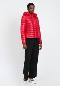 Rød puffjakke med horisontal quiltet design, hætte, frontlynlås og sidelommer, parret med brede sorte bukser og hvide hæle.