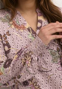 Blusa floral de tela lila claro con puños fruncidos. Presenta patrones de flores coloridos, detalles de fruncido y un diseño de escote en V.