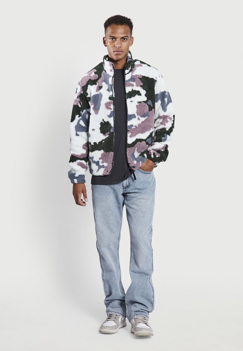 Junger Mann steht mit einer Hand in der Tasche, trägt eine camouflagemusterte Fleecejacke, ein schwarzes T-Shirt, hellblaue Jeans und weiße Sneakers vor einem weißen Hintergrund.