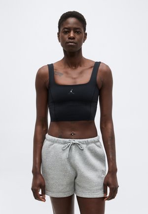Femme portant un crop top noir avec un logo et un short gris clair à cordon, debout devant un fond blanc uni.