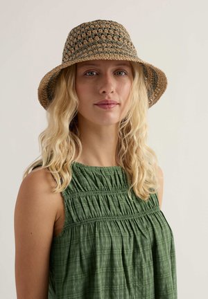 Femeie blondă cu păr ondulat, purtând un top verde fără mâneci și o pălărie largă țesută, privindu-se înainte, pe un fundal simplu.