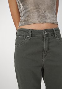 Jeans de mezclilla verde oscuro con un diseño ajustado. Incluye cinco bolsillos, pasadores para cinturón y costuras sutiles. Llevados con una blusa ligera y estampada.