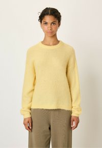 Rumena, puhasta pletena jopica s okroglim izrezom in dolgimi rokavi. Mehka tekstura, rahlo skrajšan rob, v kombinaciji s svetlo bež širokimi hlačami.