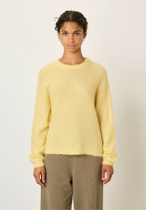 Pull jaune, en tricot duveteux, avec un col rond et des manches longues. Texture douce, ourlet légèrement court, associé à un pantalon large beige.