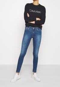Sudadera negra con el logo de "Calvin Klein", combinada con jeans ajustados azules y zapatillas blancas. El conjunto presenta un diseño casual y ajustado.