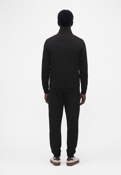 Homme vu de dos portant un haut noir à manches longues, un pantalon noir et des baskets blanches avec des semelles marron sur fond blanc.