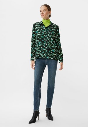 Schwarz und grün gemusterte Bluse mit langen Ärmeln, kombiniert mit blauen Skinny-Jeans und schwarzen spitzen Knöchelstiefeln. Glatte Stofftextur.